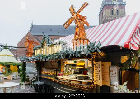 Stalle festive décorée de guirlandes saupoudrées de neige et d'un moulin à vent en bois brillant, servant nourriture et boissons dans un marché de Noël. Riga, Lettonie Banque D'Images