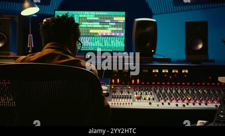 Ingénieur audio afro-américain utilisant la console de mixage sonore pour la gestion des pistes, la post-production musicale. Producteur appuyant sur les boutons de commande motorisés, regarde le logiciel d'édition. Caméra B. Banque D'Images