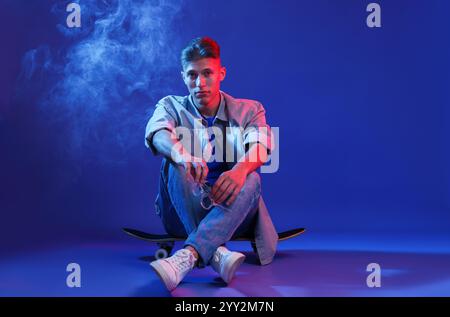 Bel homme assis sur le skateboard dans la fumée et les lumières de couleur sur fond bleu Banque D'Images
