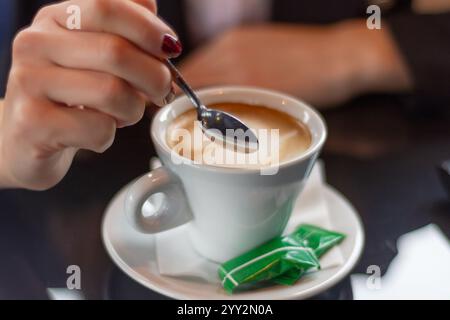 Main femelle remuant le café avec une petite cuillère en métal sur un bureau de restaurant. Concept de boisson, gros plan, mise au point sélective. Banque D'Images