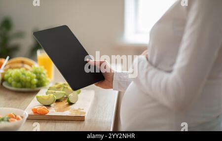 Femme enceinte utilisant une tablette pour planifier des repas sains dans une cuisine bien éclairée Banque D'Images