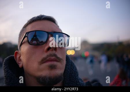 Un jeune homme portant des lunettes de soleil regarde la Tour Eiffel, dont le reflet est visible dans les verres de ses lunettes. Banque D'Images