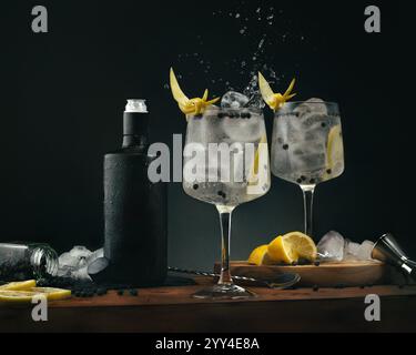 Deux verres à gin et tonic avec des garnitures de zeste de citron, entourés d'une bouteille noire, de glaçons, de citrons et de baies de genièvre sur une surface en bois Un sophis Banque D'Images