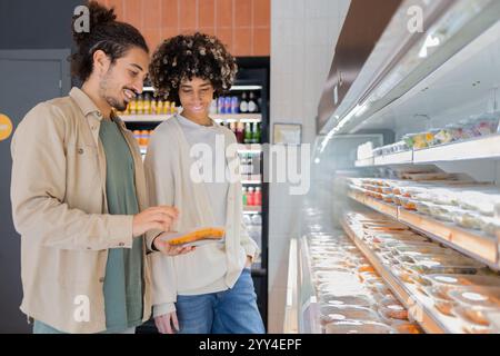 Les magasins de couple pour les repas pré-emballés dans une épicerie moderne, ils examinent un conteneur de près, souriant comme ils discutent de leur choix les étagères affichent un Banque D'Images