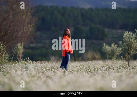 Une femme dans un pull orange vif regarde soigneusement tout en se tenant debout dans un vaste champ de fleurs blanches délicates, encadrées par des collines vertes lointaines et un c Banque D'Images