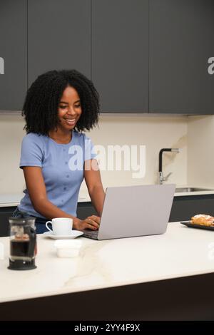Une jeune femme afro-américaine utilisant un ordinateur portable à la maison, portant une chemise bleu clair Une tasse de café et croissant sont sur le comptoir de la cuisine, créant un Banque D'Images