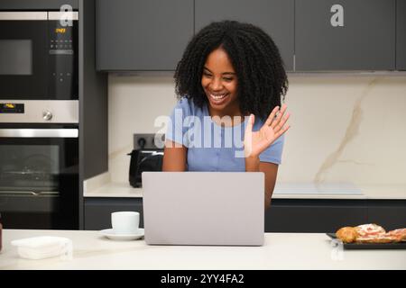 Souriant jeune femme noire ondule pendant un appel vidéo sur son ordinateur portable, assis dans une cuisine élégante Une tasse et des pâtisseries sont sur le comptoir, ajoutant de la chaleur t Banque D'Images