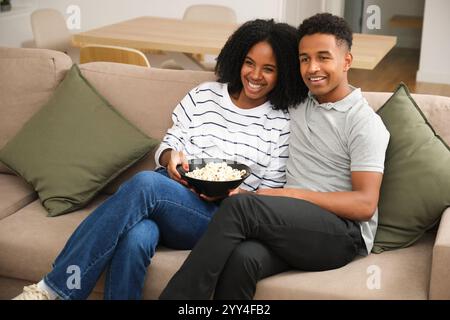 Un jeune couple noir joyeux est assis sur un canapé à la maison, profitant d'un bol de pop-corn leurs sourires reflètent le confort et le bonheur, capturant un a chaud et détendu Banque D'Images