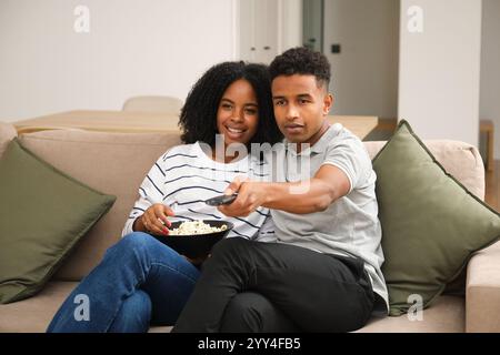 Un jeune couple noir s'assoit confortablement sur un canapé à la maison, partageant un bol de pop-corn et regardant la télévision, ils semblent détendus et joyeux dans une vie confortable ro Banque D'Images