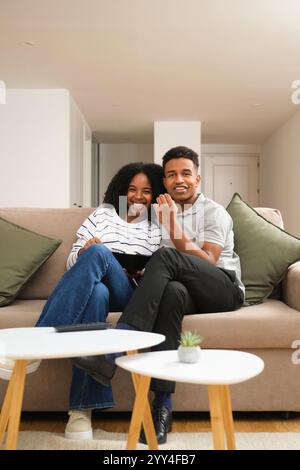 Un jeune couple afro-américain se détend sur le canapé de leur salon, souriant et profitant de moments de qualité ensemble dans un cadre confortable à la maison Banque D'Images