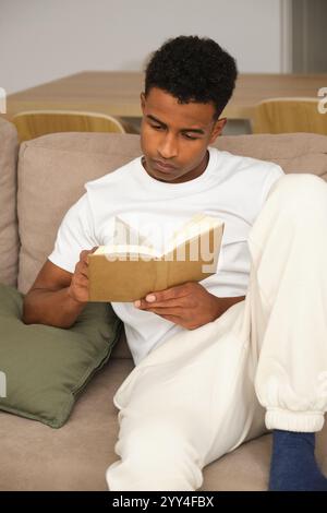 Un jeune homme afro-américain se détend à la maison, lit un livre sur le canapé, incarnant le confort et les loisirs Un instantané confortable de la vie quotidienne et de la détente Banque D'Images