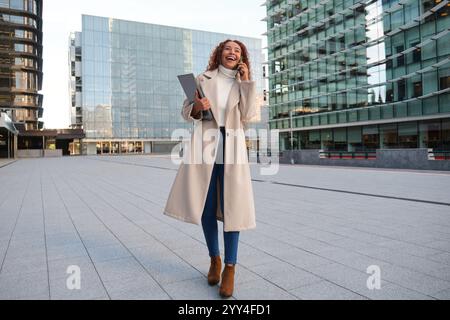 Une femme latine confiante avec des cheveux bouclés, portant un long manteau, aime un appel téléphonique elle se tient dans un paysage urbain moderne, exsudant le dynamisme et le style Banque D'Images