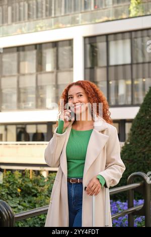 Une femme latine souriante aux cheveux bouclés parle sur son téléphone tout en se promenant dans un cadre urbain elle porte un top vert décontracté et un manteau beige, incarnant JO Banque D'Images