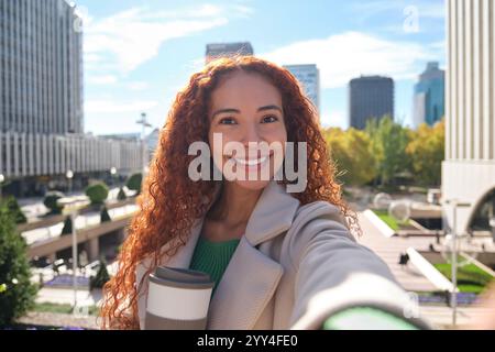 Une femme latine joyeuse aux cheveux bouclés prend un selfie dans un environnement urbain animé, tenant une tasse de café et souriant brillamment sur fond de Banque D'Images