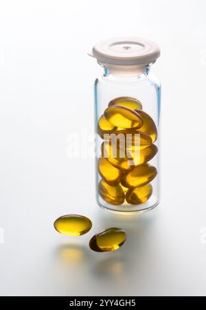 Un flacon en verre rempli de capsules jaunes se tient sur une surface réfléchissante blanche deux capsules sont placées à côté, soulignant leur texture brillante Banque D'Images