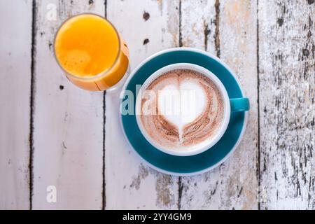 Une scène confortable avec une tasse de cappuccino avec latte art en forme de cœur, associé à un verre de jus d'orange frais, sur une table rustique en bois. Banque D'Images