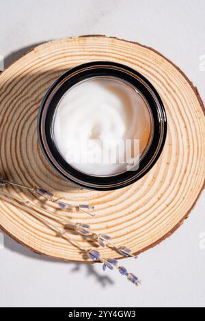 Une crème hydratante dans un bocal en verre noir se trouve sur un dessous de verre circulaire en bois, avec des brins de lavande à côté. Idéal pour la détente et la beauté sur le thème co Banque D'Images