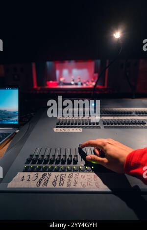 Un ingénieur du son dans une cabine de théâtre ajuste un panneau de commande pendant un spectacle. La scène montre la scène en arrière-plan, en mettant en évidence le t Banque D'Images