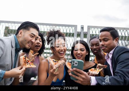 Un groupe d'amis ayant un grand temps à une fête sur le toit dans la ville, ils mangent de la pizza et rient tout en regardant quelque chose sur un smartphone, c Banque D'Images