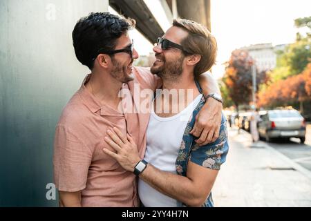 Heureux couple masculin gay embrassant et riant dans une rue animée de la ville. Un homme en chemise rose, l'autre en chemise bleue florale Banque D'Images