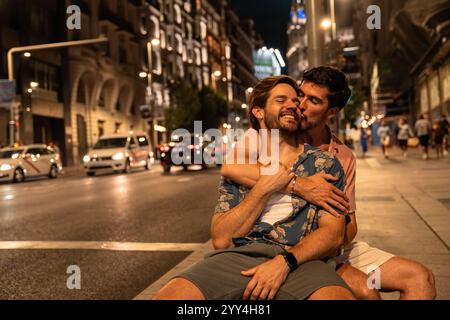 Un couple gay partage un moment tendre dans une rue animée de la ville la nuit, entouré par les lumières chaudes de la ville et la toile de fond animée de la vie urbaine tout en étant assis Banque D'Images