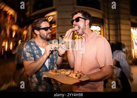 Une image joyeuse de deux hommes portant des lunettes de soleil partageant des tranches de pizza dans une rue animée de la ville, éclairée par des néons et des flashs Banque D'Images