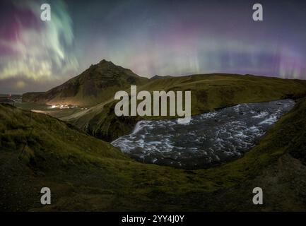 Les aurores boréales dansent au-dessus de la région de Skogafoss, avec des couleurs vives illuminant le ciel nocturne Un ruisseau coule à travers des collines moussues, créant un pois Banque D'Images