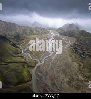 Vue aérienne des hauts plateaux islandais avec des rivières tressées qui traversent des montagnes verdoyantes sous un ciel spectaculaire et orageux, mettant en valeur la beauté brute de Banque D'Images