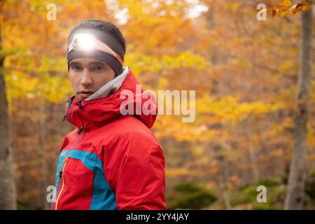 Un jeune homme dans une veste rouge avec une lampe frontale regarde vers la caméra, sur fond de forêt automnale vibrante. Il semble prêt et concentré, R Banque D'Images