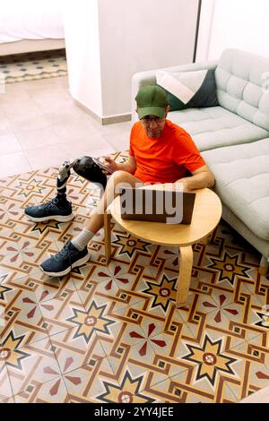 Un homme avec une jambe prothétique est assis sur un sol décoratif de tuiles, portant des vêtements de sport décontractés et une casquette, travaillant sur un ordinateur portable avec un téléphone à la main par un s confortable Banque D'Images