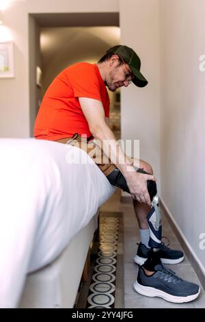 Un homme portant une casquette et une chemise rouge est assis sur un lit tout en ajustant sa jambe prothétique le cadre est une pièce confortable et bien éclairée, illustrant la vie quotidienne an Banque D'Images