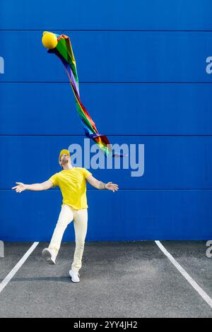 Une personne vêtue de jaune vole joyeusement un cerf-volant coloré contre un mur bleu frappant. La scène vibrante capture un moment ludique de plaisir en plein air et Banque D'Images