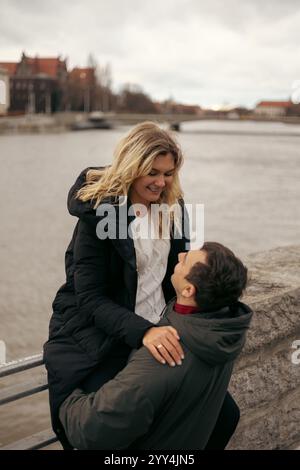 Un jeune couple marié, partage un moment de joie en marchant le long de la rivière à Wroclaw, en Pologne pendant la saison fraîche. La femme est assise sur un RA Banque D'Images