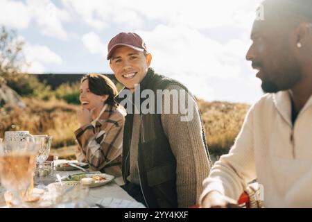Portrait de jeune homme souriant portant un chapeau assis à table avec des amis pour dîner le jour ensoleillé Banque D'Images