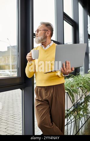 L'homme mûr avec une barbe se tient près d'une grande fenêtre, sirotant du café et utilisant un ordinateur portable. Banque D'Images