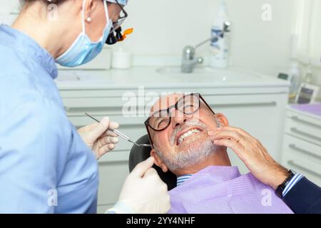 Dentiste portant une loupe et un masque chirurgical examinant un patient âgé souffrant de maux de dents dans une clinique dentaire brillante Banque D'Images