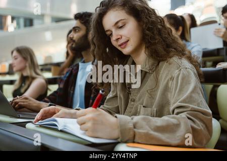 Jeune femme aux cheveux bouclés écrivant dans un livre alors qu'elle était assise dans une salle de classe à l'université Banque D'Images