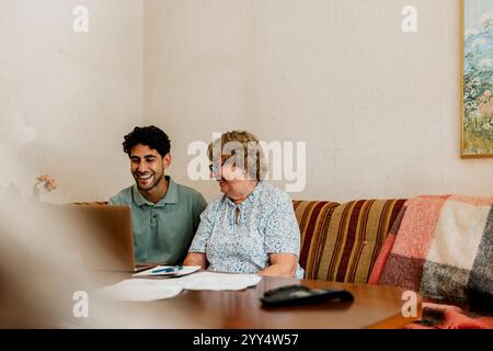 Infirmière masculine souriante utilisant un ordinateur portable tout en étant assis avec une femme âgée sur le canapé à la maison Banque D'Images