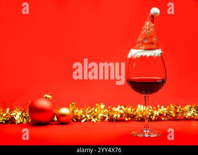 Un verre de vin rouge surmonté d'un chapeau de Père Noël, placé sur une surface rouge vif avec des ornements de Noël et des guirlandes dorées en arrière-plan, évoquant un fès Banque D'Images