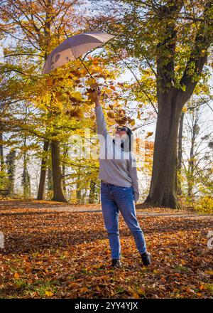 Femme se tient dans un parc d'automne et jette des feuilles d'or d'un parapluie à elle-même, souriant. Vokruz feuilles jaunes vives, créant un a chaud et ludique Banque D'Images