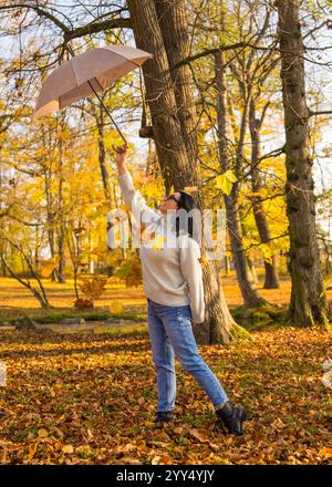 Femme se tient dans un parc d'automne et jette des feuilles d'or d'un parapluie à elle-même, souriant. Vokruz feuilles jaunes vives, créant un a chaud et ludique Banque D'Images