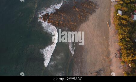 Vue aérienne d'une plage tropicale au coucher du soleil avec des palmiers, rivage rocheux, et les visiteurs dispersés appréciant le paysage côtier serein. Banque D'Images