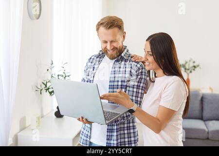 Jeune couple heureux debout dans un salon confortable regardant l'écran d'ordinateur portable, regardant la vidéo, achetant Banque D'Images