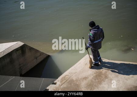 L'eau orange sale de la canalisation d'égout se mélange avec l'eau de la rivière dans la ville. Pollution de l'environnement, Un baril toxique flotte, observe le travailleur. Banque D'Images