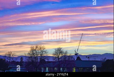 Le crépuscule tombe sur un quartier de Denver avec un ciel de coucher de soleil rayé de couleurs vives. Une grande grue et les montagnes voisines sont visibles en arrière-plan. Banque D'Images