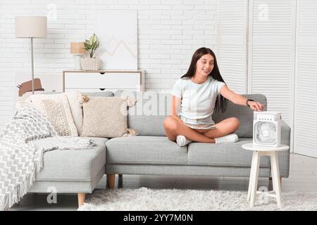 Belle jeune femme lisant magazine près du ventilateur de table portable avec humidificateur dans le salon Banque D'Images