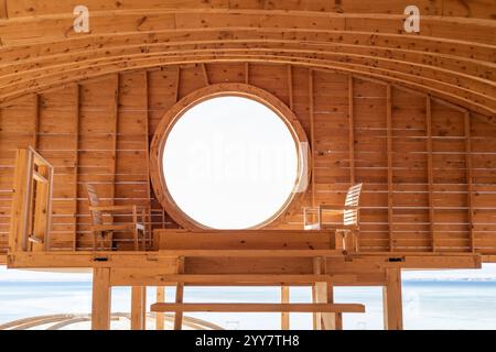 Cette image accueillante présente un intérieur en bois serein avec une fenêtre circulaire unique, offrant une vue imprenable sur l'océan Banque D'Images