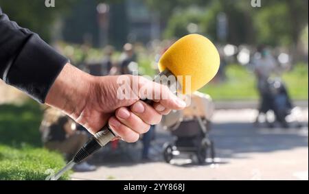 Main dans un pull gris tenant un microphone à main en métal avec pare-brise en mousse jaune pour interviewer à l'extérieur dans un parc Banque D'Images