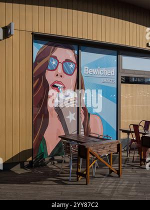 Façade de Bewiched, une chaîne locale indépendante de cafés, Rushden Lakes, Northamptonshire, Royaume-Uni ; avec peinture murale représentant une femme buvant du café mousseux Banque D'Images