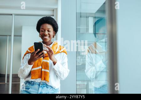 Une femme architecte confiante dans une chemise blanche et un pull orange, debout dans un environnement de bureau élégant, s'engageant avec son smartphone. Son exp joyeux Banque D'Images